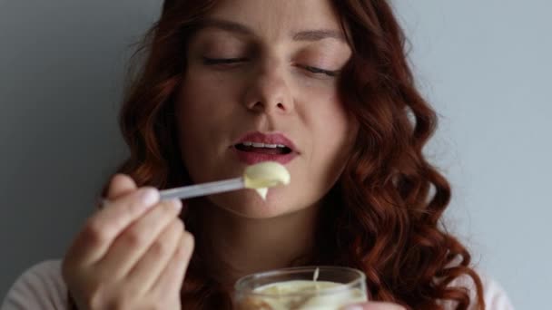 Позитивна кучерява біла дівчина їсть смачний вершковий десерт, насолоджуючись смаком на сірому фоні. Немає дієти
 - Кадри, відео
