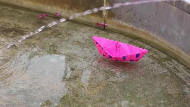 Маленький мальчик кладёт ярко-розовую бумажную лодку в воду и отталкивает её. Дети веселятся у воды. - Кадры, видео