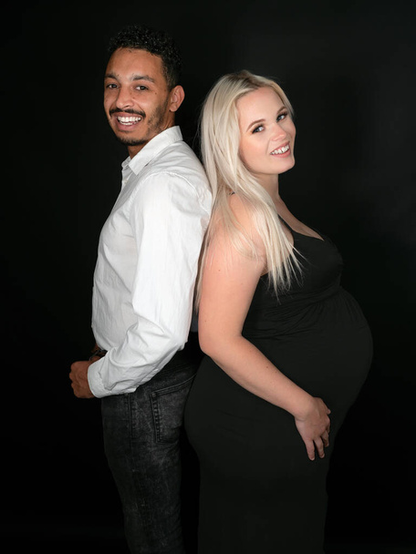 Vista del perfil de una pareja multiétnica elegante y feliz que espera un bebé en una pose espalda con espalda - Foto, imagen