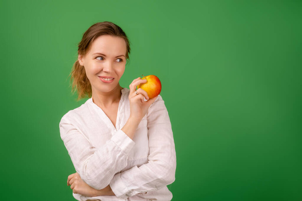 A good-looking woman of forty in a white shirt on a green background smiles and holds a fresh apple in her hands. Healthy eating, vegetarianism, healthy appearance concept. - Foto, Imagen