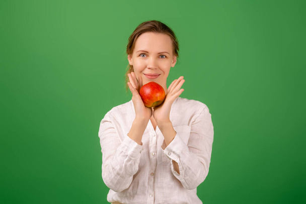 Kaunis keski-ikäinen nainen valkoisessa paidassa pitää omenaa kädessään vihreällä taustalla. Terveellinen syöminen, kasvissyönti, terveellinen ulkonäkö käsite. - Valokuva, kuva