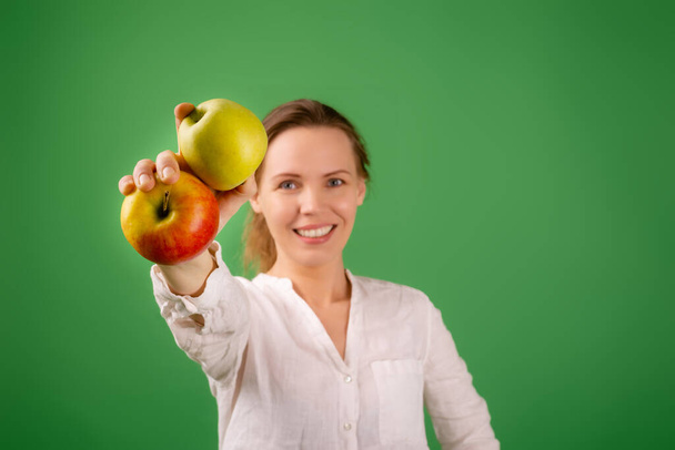 A beautiful middle-aged woman in a white shirt shows apples on a green background. The concept of diet, healthy food, vegetarianism. - Photo, image