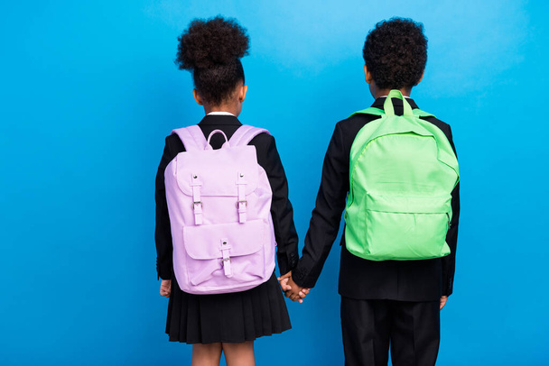 Back rear spine view photo of dark skin boy and girl hold hands wear rucksack isolated on blue color background - Valokuva, kuva