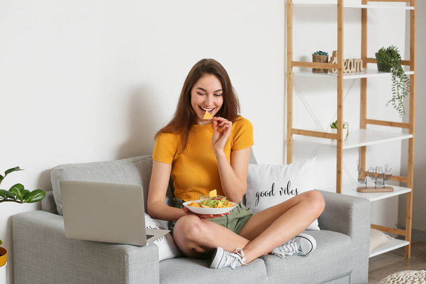 Beautiful young woman with laptop eating tasty nachos at home - Фото, зображення