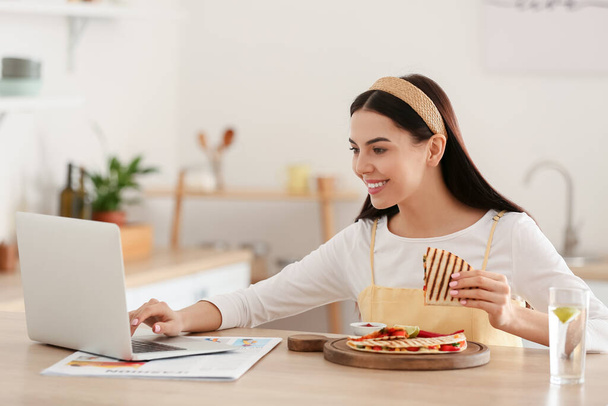 Όμορφη νεαρή γυναίκα με φορητό υπολογιστή τρώει νόστιμο quesadilla στο σπίτι - Φωτογραφία, εικόνα