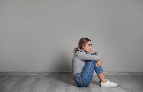 Καταθλιπτική νεαρή γυναίκα που κάθεται στο πάτωμα κοντά στον γκρίζο τοίχο - Φωτογραφία, εικόνα