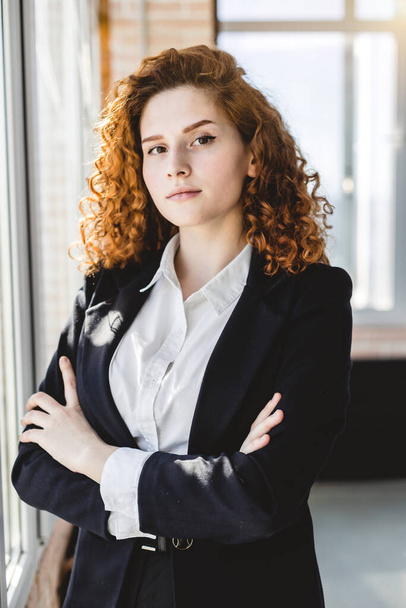 Portrait of a beautiful successful young business woman with red curly hair in the office interior stands near the window - Фото, зображення