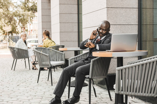 Portrait of a black African American businessman in a suit sitting in a city cafe outdoors and talking on the phone. - Foto, Imagem
