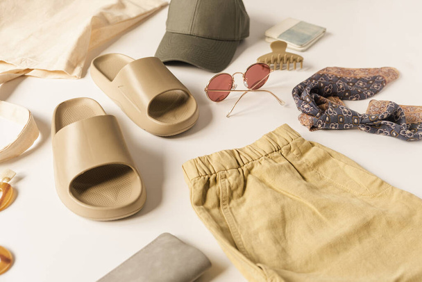 Ουδέτερο σκονισμένο μπεζ χρώμα γυναικεία ρούχα μόδας και αξεσουάρ για καλοκαιρινές διακοπές παραλία chill. Καπελάκι, παντόφλες, γυαλιά ηλίου, μπαρέτα, σορτς, τσάντα για ψώνια σε λευκό φόντο - Φωτογραφία, εικόνα