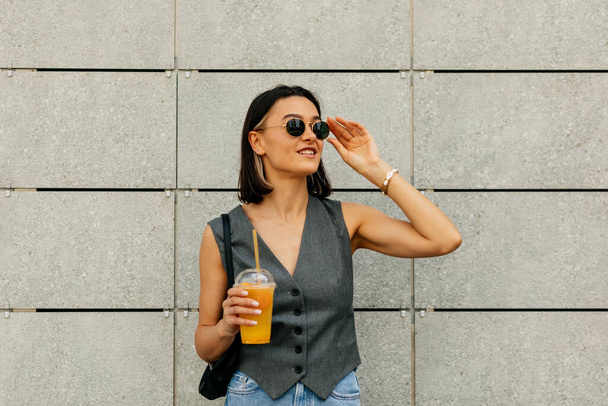 Blithesome Avrupalı kadın sokak arka planında gözlüklerine dokunuyor. Harika esmer kadın model, gri duvarda meyve suyu var. - Fotoğraf, Görsel