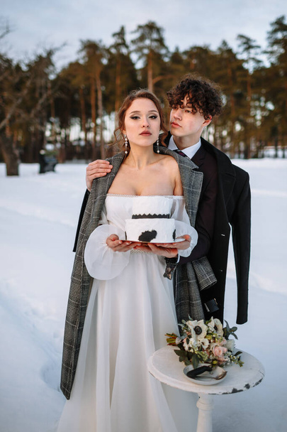 The bride and groom hold a wedding cake in their hands, standing in the snow in the forest. - Fotoğraf, Görsel