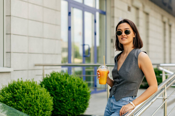 Αξιολάτρευτο υπέροχο κορίτσι με γυαλιά ηλίου και γιλέκο και τζιν να περπατά έξω και να πίνει χυμό στην πόλη - Φωτογραφία, εικόνα