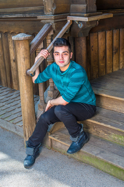 In einem grün gestreiften langärmeligen Henley-Hemd, schwarzer Hose und Lederstiefeln sitzt ein junger, gutaussehender Mann auf der Treppe vor einem Holzhaus und macht eine Pause.. - Foto, Bild