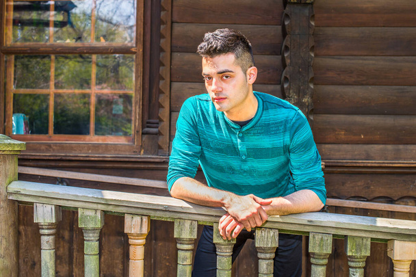 In einem grün gestreiften, langärmeligen Henley-Hemd beugt sich ein junger, gutaussehender Mann auf einem Geländer vor einem Holzhaus und entspannt - Foto, Bild