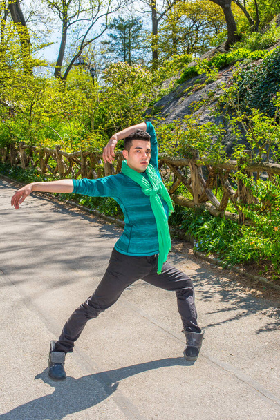 Habillé d'une chemise verte à manches longues, d'un foulard, d'un pantalon noir et de bottes en cuir, étirant les bras et retournant les jambes, un jeune homme fait de l'exercice à l'extérieur au printemps - Photo, image
