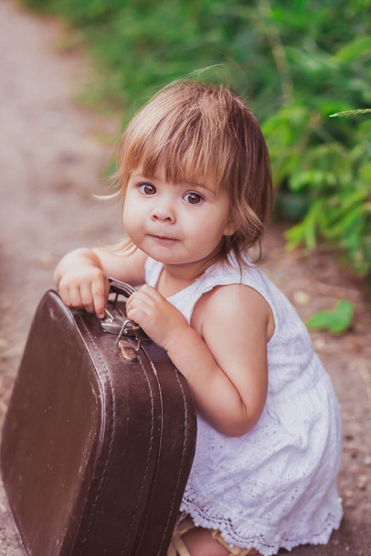 Charming baby in a white sundress tries to open an old suitcase - Foto, afbeelding
