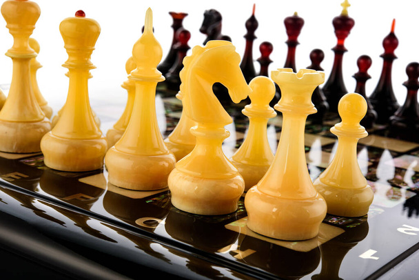 Φυσικό κεχριμπάρι διαφορετικά κομμάτια σκακιού που στέκεται σε μαργαριτάρι πίνακα σε λευκό. Close up παιχνίδι έννοια ανταγωνισμού, Κλασικό Τουρνουά της αντιπαράθεσης. - Φωτογραφία, εικόνα