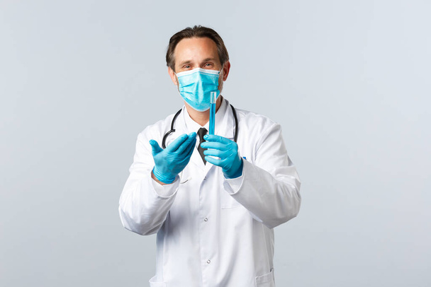 Covid-19, preventing virus, healthcare workers and vaccination concept. Satisfied smiling doctor in medical mask and gloves pointing at test-tube with vaccine sample, pleased with good result - Foto, afbeelding