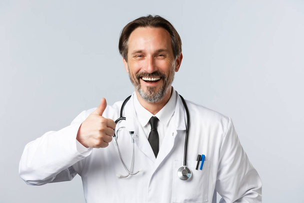 Covid-19, preventing virus, healthcare workers and vaccination concept. Cheerful middle-aged male doctor in white coat laughing happy and thumb-up in approval, recommend product or services - Photo, Image