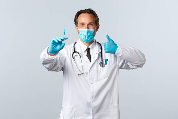 Covid-19, zapobieganie wirusom, pracownikom służby zdrowia i koncepcji szczepień. Pewny siebie lekarz w masce medycznej i rękawiczkach kciuki do góry, zalecamy szczepionkę z koronawirusu, trzymając strzykawkę - Zdjęcie, obraz