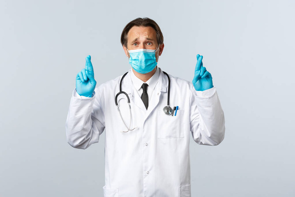Covid-19, preventing virus, healthcare workers and vaccination concept. Troubled hopeful doctor in medical mask and gloves, cross fingers good luck, praying or making wish, white background - Фото, изображение