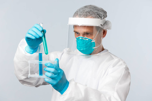 Covid-19, preventing virus, healthcare workers and vaccination concept. Tech lab doctor in clinic wearing personal protective equipment working over vaccine, looking at test-tubes samples - Photo, Image