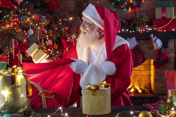 Santa Claus with a bag with presents gifts near fireplace and Christmas tree. Festive interior inside wooden house, New Year's cheerful mood Spirit of Christmas. Senior man with real white beard cosplay Father Christmas. - Fotó, kép