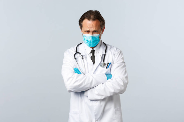 Covid-19, preventing virus, healthcare workers and vaccination concept. Offended timid doctor in medical mask and gloves looking insulted and upset, cross hands chest and frowning gloomy - Photo, Image