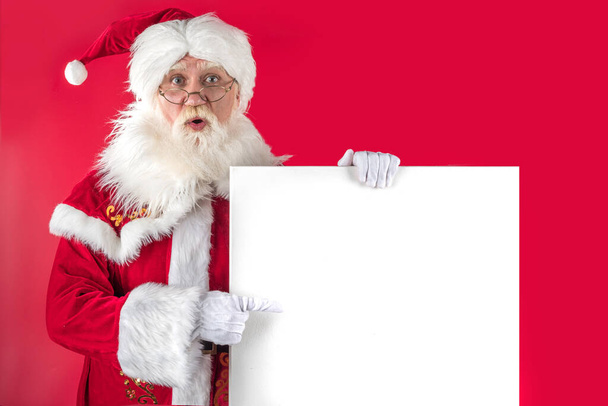 Xmas Santa Claus pointing finger, holding whiteboard on red background. Emotional senior male model old man with a natural white beard Father Christmas. Joyful character for advertising - Foto, Imagem