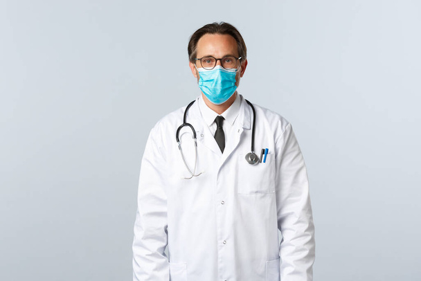 Covid-19, prevención de virus, trabajadores sanitarios y concepto de vacunación. Médico masculino serio confiado en bata blanca y máscara médica, usando gafas, paciente listo del tratamiento con la gripe - Foto, Imagen