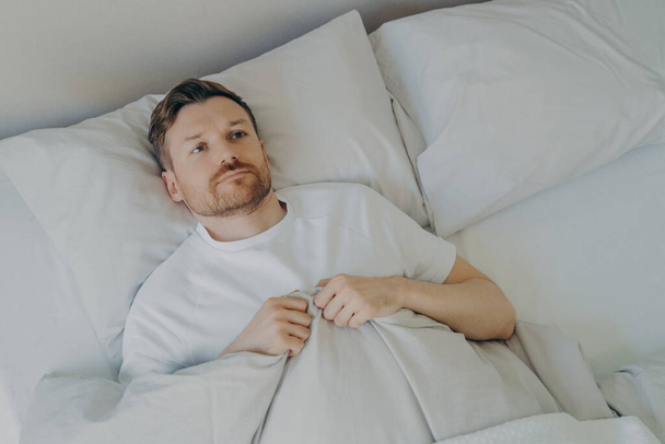 Μοναχικός αναστατωμένος νεαρός γενειοφόρος ξαπλωμένος στο κρεβάτι με ανοιχτά μάτια - Φωτογραφία, εικόνα
