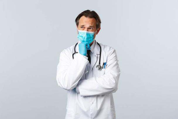 Covid-19, preventing virus, healthcare workers and vaccination concept. Thoughtful serious male doctor in medical mask and gloves thinking, making choice or decision, white background - Photo, Image
