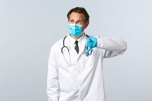 Covid-19, zapobieganie wirusom, pracownikom służby zdrowia i koncepcji szczepień. Zdenerwowany mężczyzna lekarz w masce medycznej, rękawiczki pokazują kciuk w dół i patrząc w lewym górnym rogu promo - Zdjęcie, obraz