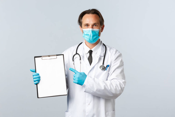 Covid-19, preventing virus, healthcare workers and vaccination concept. Professional doctor at clinic pointing at clipboard paper, explain disease or showing chart, wear medical mask and gloves - Foto, afbeelding
