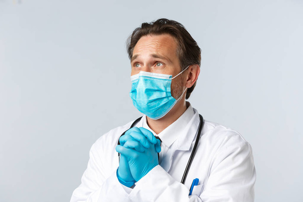 Covid-19, preventing virus, healthcare and vaccination concept. Close-up of desperate hopeful doctor look concerned, praying. Therapist in medical mask and gloves clench hands in pleading gesture - Фото, изображение