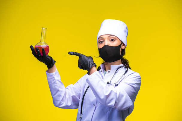 Молодая медсестра в маске для лица и рук держит химическую фляжку и указывает на нее пальцем. Высокое качество фото - Фото, изображение