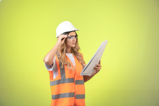 Ženská inženýrka v helmě a výstroji, držící projektový plán a s brýlemi. Kvalitní fotografie - Fotografie, Obrázek