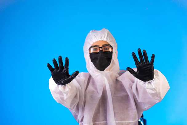 Özel önleyici üniformalı ve siyah maskeli dezenfektan mavi arka planda dur işareti yapıyor. Yüksek kalite fotoğraf - Fotoğraf, Görsel