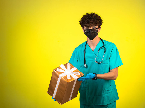 Σγουρά μαλλιά αγόρι σε ιατρική στολή και το χέρι, μάσκες προσώπου κρατώντας ένα κουτί δώρου. Υψηλής ποιότητας φωτογραφία - Φωτογραφία, εικόνα