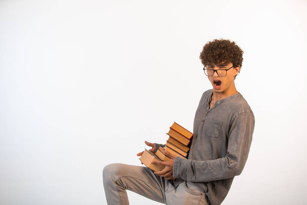 Chłopak z kręconymi włosami, noszący optymistyczne okulary, ledwo trzyma na kolanach zapasy książek. Wysokiej jakości zdjęcie - Zdjęcie, obraz