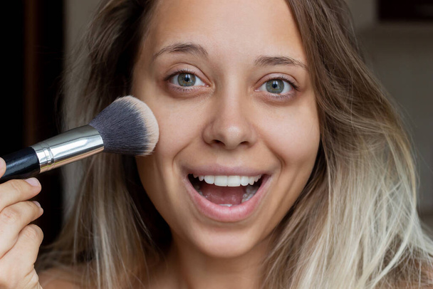 Close-up of a young caucasian smiling blonde woman without makeup uses a brush applying powder to the skin of the face on a dark background - Photo, image