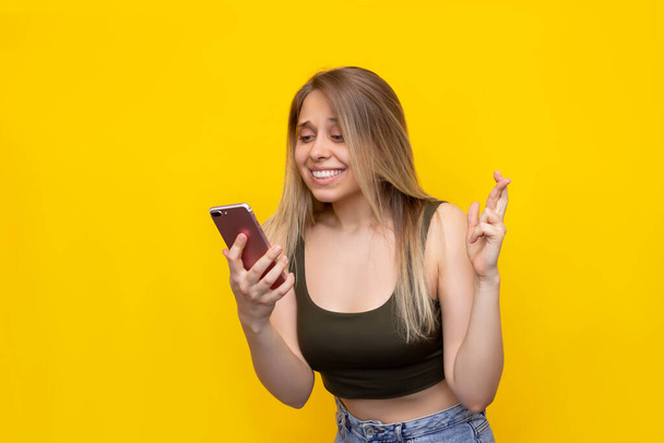 A young pretty caucasian smiling blonde woman crosses her fingers for good luck looking at the phone screen waiting for the results of the lottery or exams isolated on a bright color yellow background - Foto, immagini