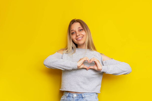 A young caucasian pretty charming blonde woman forming a heart shape with her hands isolated on a bright color yellow background. Beautiful cute girl shows her love. Charity concept  - Фото, изображение