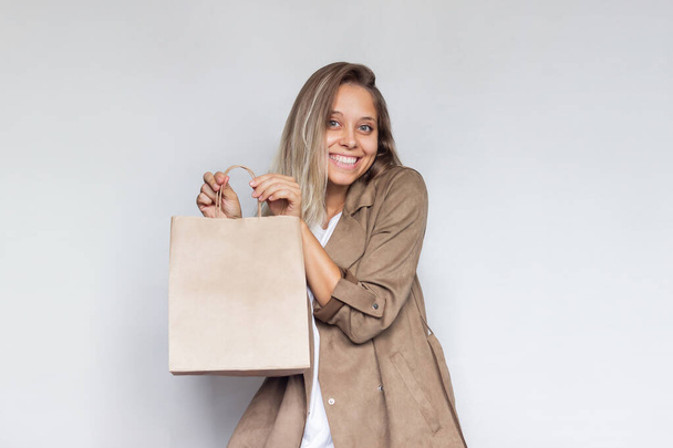 A young beautiful caucasian smiling blonde woman in a brown coat holds and demonstrates paper eco bag with copy empty space for the logo isolated on a light gray background.Shopping and fashion - Φωτογραφία, εικόνα