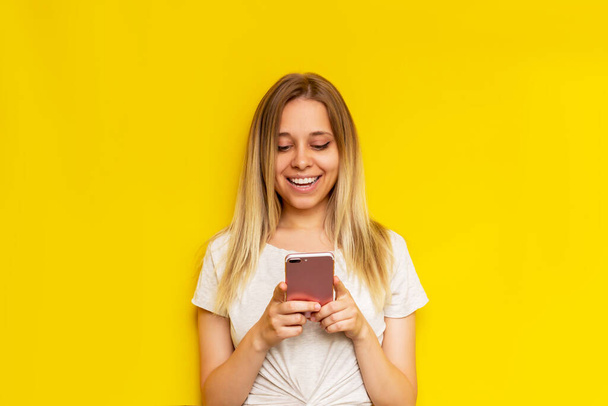 A young caucasian happy pretty blonde woman smiles and holds a mobile phone looking at the screen isolated on a bright color yellow background. The girl uses her smartphone - Fotografie, Obrázek