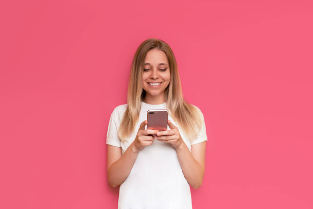 A young caucasian happy pretty blonde woman smiles and holds a mobile phone looking at the screen isolated on a bright color pink background. The girl uses her smartphone - Foto, Imagen