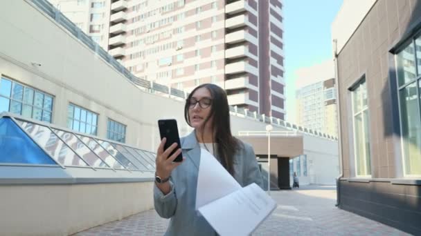 vrouw in een formeel pak praat op een video link en kijkt naar papieren lopen door de straat, slow motion - Video