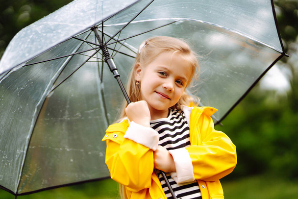 Little girl with transparent umbrella  playing in the rain on sunny autumn day. Beautiful girl  in a yellow cloak and rubber boots. Fall outdoor activity by rainy weather. - Foto, Imagem