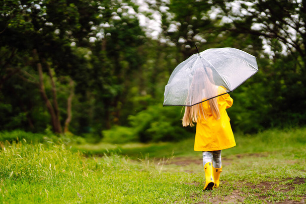Little girl with transparent umbrella  playing in the rain on sunny autumn day. Beautiful girl  in a yellow cloak and rubber boots. Fall outdoor activity by rainy weather. - Φωτογραφία, εικόνα