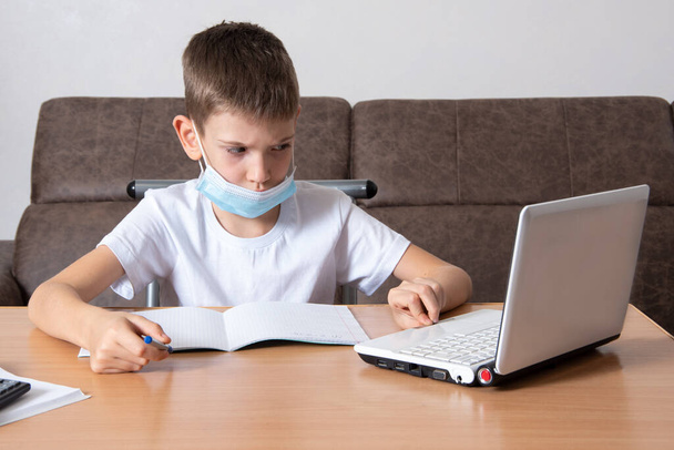 Distance learning during an epidemic - A serious schoolboy in a protective mask on his face looks at a laptop, writes information in a notebook, sitting at a table at home. Back to school concept - Фото, зображення